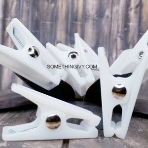 May include: Six white plastic clips with silver metal spring and button. The clips are arranged in a cluster on a wooden surface.