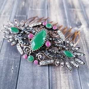 May include: A gold-tone hair comb with a decorative design featuring green and pink gemstones. The comb has a floral pattern with intricate details.