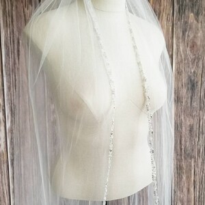 May include: A white bridal veil with a beaded trim. The veil is on a white mannequin against a wood background.