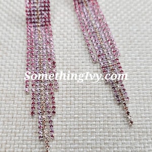 Può includere: Un paio di lunghi orecchini pendenti rosa e argento con un design a cascata di strass. Gli orecchini sono su uno sfondo di tessuto beige.