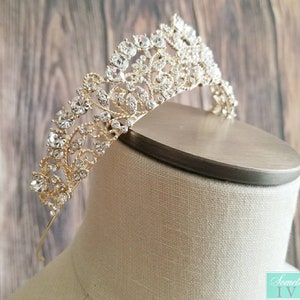 Puede incluir: Una tiara dorada con brillantes pedrer&iacute;a transparente. La tiara presenta un dise&ntilde;o floral ornamentado con m&uacute;ltiples filas de piedras. Se exhibe sobre una cabeza de maniqu&iacute;, mostrando sus intrincados detalles y su elegante estilo.