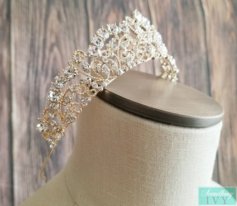 Puede incluir: Una tiara dorada con brillantes pedrer&iacute;a transparente. La tiara presenta un dise&ntilde;o floral ornamentado con m&uacute;ltiples filas de piedras. Se exhibe sobre una cabeza de maniqu&iacute;, mostrando sus intrincados detalles y su elegante estilo.