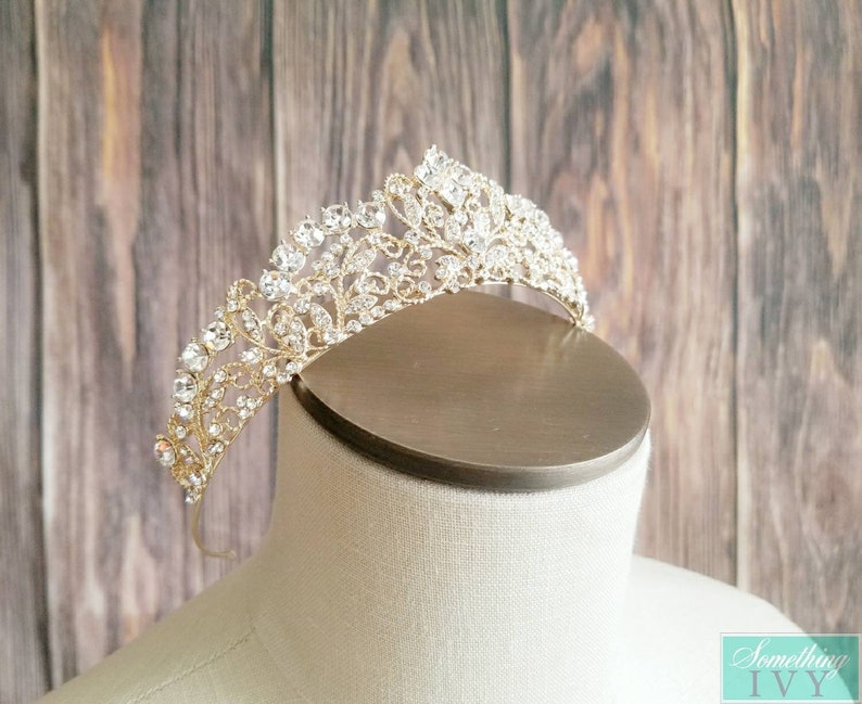 Puede incluir: Una tiara dorada con intrincados filigranas y brillantes cristales transparentes. El tocado nupcial presenta un dise&ntilde;o floral y de hojas, perfecto para bodas u ocasiones especiales. La tiara se exhibe en una cabeza de maniqu&iacute;.