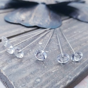 Pode incluir: Um conjunto de seis alfinetes de flores de cristal de imitação transparente com hastes prateadas. Os cristais de imitação são redondos e têm um design facetado. Os alfinetes estão dispostos em forma de leque sobre uma superfície de madeira.