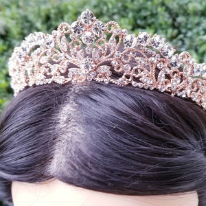 Puede incluir: Una tiara de oro rosa con detalles de cristal transparente. La corona ornamentada presenta un delicado dise&ntilde;o de filigrana y est&aacute; adornada con brillantes pedrer&iacute;a. La tiara se encuentra sobre un fondo de cabello oscuro y vegetaci&oacute;n.