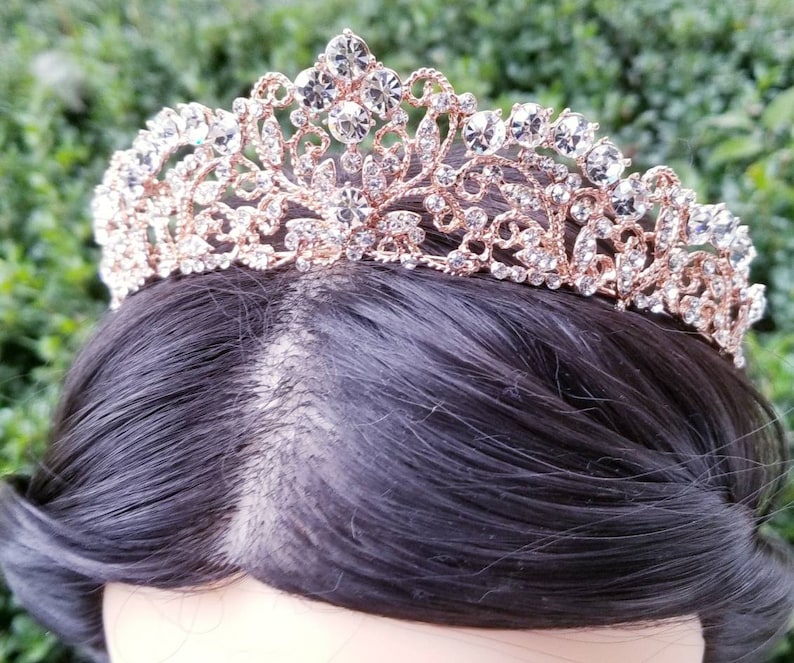 Puede incluir: Una tiara de oro rosa con detalles de cristal transparente. La corona ornamentada presenta un delicado dise&ntilde;o de filigrana y est&aacute; adornada con brillantes pedrer&iacute;a. La tiara se encuentra sobre un fondo de cabello oscuro y vegetaci&oacute;n.