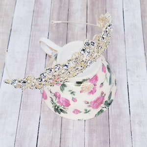 Puede incluir: Una tiara dorada adornada con brillantes pedrer&iacute;a transparente. La tiara est&aacute; colocada sobre una fina diadema plateada. La tiara descansa sobre una taza de t&eacute; floral con rosas rosas y hojas verdes.