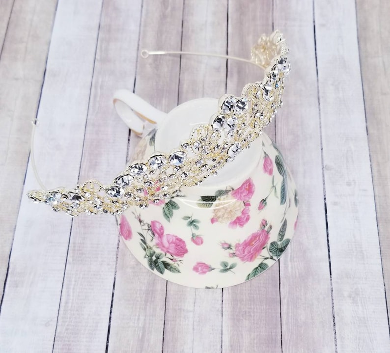 Puede incluir: Una tiara dorada adornada con brillantes pedrer&iacute;a transparente. La tiara est&aacute; colocada sobre una fina diadema plateada. La tiara descansa sobre una taza de t&eacute; floral con rosas rosas y hojas verdes.
