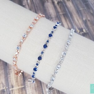 Pu&ograve; includere: Tre braccialetti delicati con accenti di cristallo trasparente e blu. I braccialetti sono su uno sfondo di tessuto bianco.