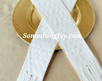 May include: A pair of white, sheer gloves with a delicate dotted pattern, resting on a gold-colored charger plate. The gloves are long, extending past the wrist. The text "SomethingIvy.com" is visible.
