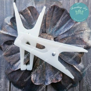 May include: Two white plastic clothespins with a metal spring. The clothespins are on a brown wooden surface with a floral design.