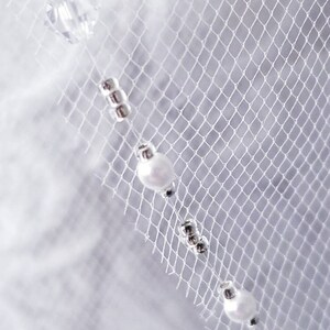 May include: White tulle fabric with a beaded trim. The trim features small silver beads and larger white pearl-like beads.