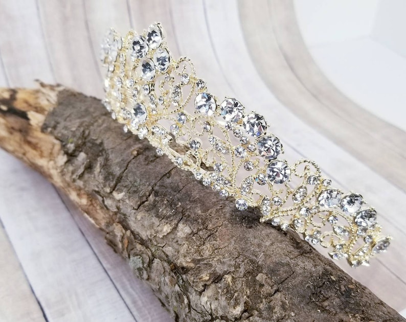 Puede incluir: Una tiara dorada adornada con brillantes pedrer&iacute;a transparente. La tiara presenta un dise&ntilde;o floral ornamentado con una serie de grandes pedrer&iacute;as redondas en la parte superior. La tiara se muestra sobre un trozo de madera.