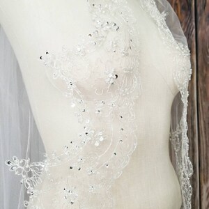 May include: A white bridal veil with a delicate lace pattern and silver embellishments. The lace features a floral design with small, sparkling beads.