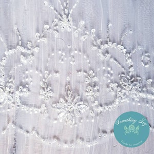 May include: White tulle fabric with a delicate beaded pattern. The fabric has a scalloped edge and is perfect for adding a touch of elegance to any project.