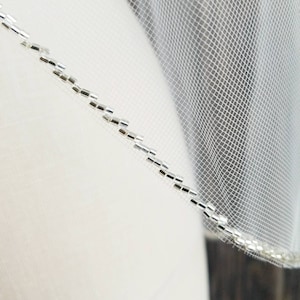 May include: A white bridal veil with a silver beaded trim. The veil is made of a fine mesh fabric and is designed for a wedding ceremony.