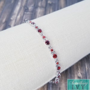 May include: A silver bracelet with a delicate chain and red gemstones. The bracelet is displayed on a white fabric surface.