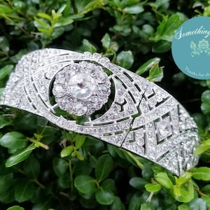 以下が含まれることがあります： 中央に円形デザインと幾何学模様があり、多数の透明なクリスタルがちりばめられたシルバーのティアラ。緑の葉を背景に、右上には円形のロゴがあります。