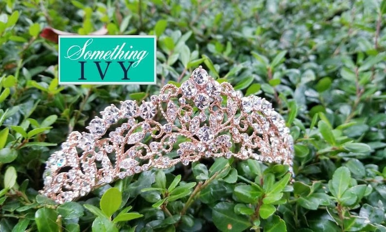 Puede incluir: Una tiara de oro rosa con detalles de cristal transparente, sobre un lecho de hojas verdes. La tiara presenta un dise&ntilde;o ornamentado con piedras brillantes. La imagen incluye un rect&aacute;ngulo turquesa con el texto "Something IVY".