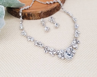 May include: A silver necklace and earrings set with clear crystal stones. The necklace features a large oval-shaped crystal in the center and smaller crystals clustered around it.
