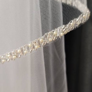 May include: White bridal veil with a delicate, beaded trim. The veil is made of a fine, sheer mesh fabric. The trim features small, round pearls and sparkling accents, adding a touch of elegance to the design.