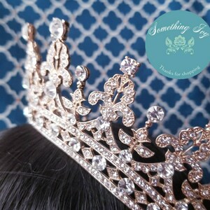 Puede incluir: Una corona dorada con brillantes transparentes. La corona tiene una punta puntiaguda y un dise&ntilde;o decorativo con motivos de flor de lis.