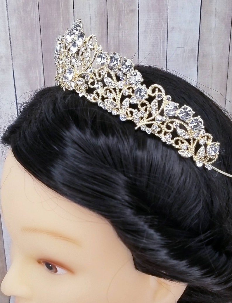 Puede incluir: Una tiara dorada adornada con brillantes y claros pedrer&iacute;a. La tiara presenta un dise&ntilde;o ornamental de filigrana con un pico central y una serie de elementos decorativos m&aacute;s peque&ntilde;os. Los brillantes est&aacute;n engastados de forma que captan la luz.