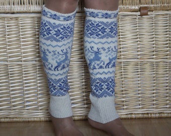 May include: A pair of white knitted leg warmers with a blue and white geometric pattern featuring reindeer. The leg warmers are worn by a person standing in front of a wicker basket.