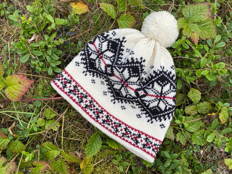 May include: White knitted hat with a black and red geometric pattern and a white pom pom on top.