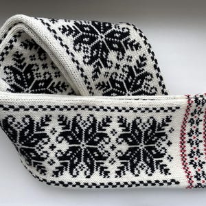 Könnte beinhalten: Ein gefalteter, weißer Strickschal mit einem schwarzen geometrischen Muster. Der Schal hat ein sich wiederholendes schneeflockenartiges Design und einen Rand mit roten Akzenten. Der Schal ist aus einem weichen, warmen Material gefertigt.