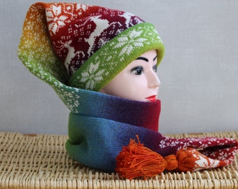 May include: A colorful knitted hat with a long scarf. The hat has a rainbow gradient and a white snowflake pattern. The scarf is a gradient of blue, green, purple, and red with a white snowflake pattern. The scarf has a large orange pom-pom.