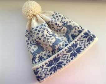 May include: White knitted beanie with a blue and white patterned design featuring reindeer and snowflakes. The beanie has a white pompom on top.