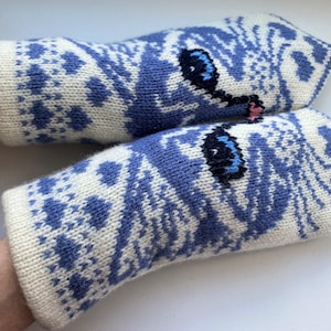 Könnte beinhalten: Ein Paar gestrickte Fäustlinge in Creme und Blau. Die Fäustlinge haben ein sich wiederholendes geometrisches Muster und ein Katzengesicht-Design auf der Rückseite jeder Hand. Die Augen der Katze sind blau und die Nase rosa.