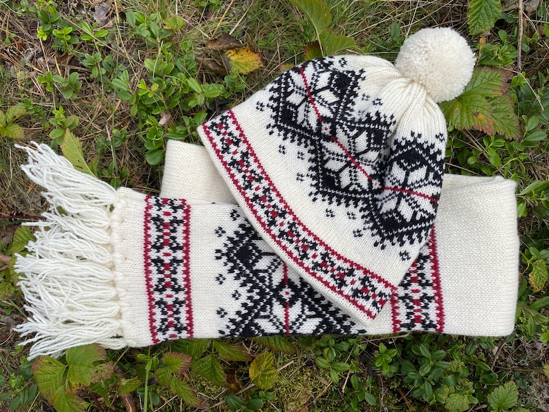 May include: White knitted hat and scarf set with a black and red geometric pattern. The hat has a white pompom on top.