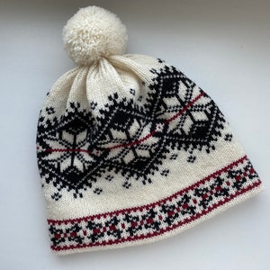 May include: White knitted hat with a black and red geometric pattern and a white pom pom on top.