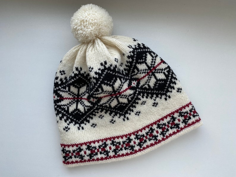 May include: White knitted hat with a black and red geometric pattern and a white pom pom on top.