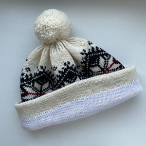 May include: White knit beanie with a black and red geometric pattern and a white pom pom on top.