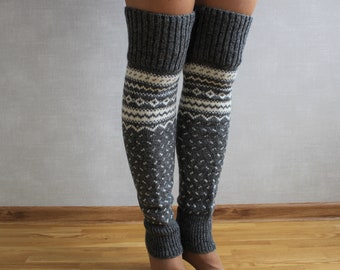May include: A pair of gray and white knit leg warmers. The leg warmers feature a ribbed cuff and a patterned design in white and gray. The design includes geometric shapes and a diamond pattern. The leg warmers are knee-high.
