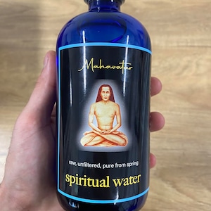 May include: A blue glass bottle with a black label that says "Mahavatar" and "spiritual water". The label also features a drawing of a person sitting in a meditative pose. The text on the label also says "raw, unfiltered, pure from spring".