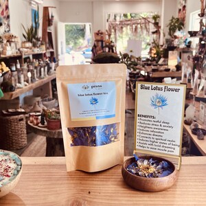 May include: A brown paper bag with a label that reads "prana blue lotus flower tea" and a picture of a blue lotus flower. The bag contains dried blue lotus flower petals. A sign next to the bag reads "Blue Lotus Flower" and lists the benefits of blue lotus flower tea, including promoting restful sleep, reducing stress and anxiety, and enhancing dreams.