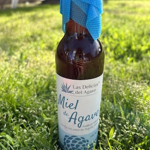 May include: A glass bottle of agave honey with a blue cloth covering the top. The label reads "Las Delicias del Agave Miel de Agave Producto 100% natural Hecho con amor en Tequila, Jalisco".