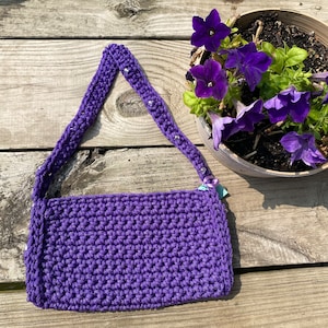 May include: A purple crocheted handbag with a beaded strap. The bag is sitting on a wooden surface.