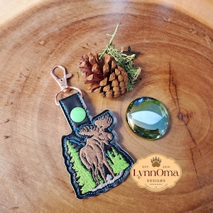 May include: A brown moose with antlers embroidered on a black leather keychain. The keychain has a green circle and a silver metal clasp. The keychain is on a wooden surface with a pine cone and a green glass marble.