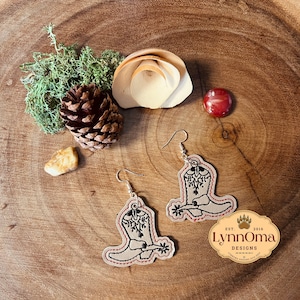 May include: A pair of brown leather earrings with a black embroidered cowboy boot design. The earrings are hanging from silver hooks. The earrings are labeled "LynnOma Designs" with the text "Est. 2016".