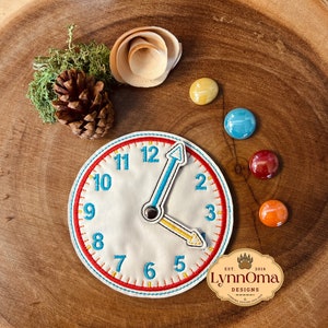 Puede incluir: Un reloj de fieltro blanco con costuras rojas, naranjas y azules. La esfera del reloj tiene números negros y una manecilla de hora azul y una manecilla de minutos amarilla. El reloj está sobre una superficie de madera con piedras decorativas de colores.