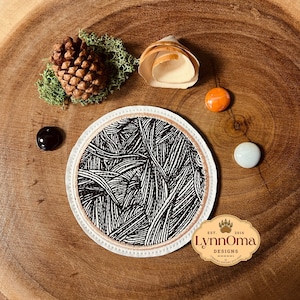 May include: A round coaster with a black and white abstract design, featuring a white border and a tan trim. The coaster is surrounded by decorative elements on a wooden surface, including a pinecone, moss, and small stones. The coaster has the text "LynnOma Designs".