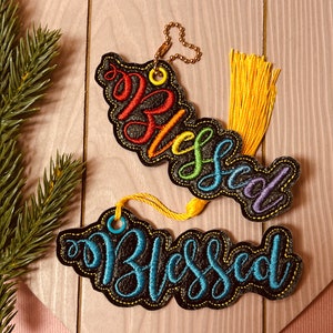 Puede incluir: Dos adornos de fieltro negro con la palabra "Blessed" bordada en hilo de colores. El adorno superior tiene hilo de colores arco&iacute;ris y el adorno inferior tiene hilo azul. Ambos adornos tienen un lazo para colgar.