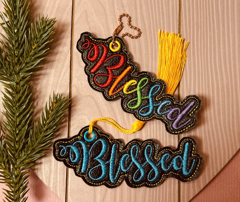 Puede incluir: Dos adornos de fieltro negro con la palabra "Blessed" bordada en hilo de colores. El adorno superior tiene hilo de colores arco&iacute;ris y el adorno inferior tiene hilo azul. Ambos adornos tienen un lazo para colgar.