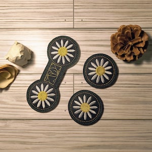 May include: An overhead shot displays three daisy-themed patches and a connecting piece, all with black backgrounds, white petals, and yellow centers. A pinecone and two wooden decorative pieces are also present on a light wood surface.