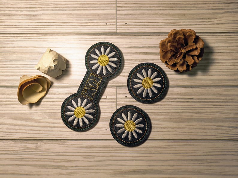 May include: An overhead shot displays three daisy-themed patches and a connecting piece, all with black backgrounds, white petals, and yellow centers. A pinecone and two wooden decorative pieces are also present on a light wood surface.